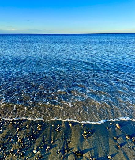 Photo of view looking out at the open ocean from the shore.  The water is nearly flat with just small ripples on it.  Foam from the gentle surf is seen in the foreground.  The water is blue, reflecting the solid blue sky.  The horizon is slightly orange from the sun setting in the background.

Taken on a beach in Southeastern Massachusetts.