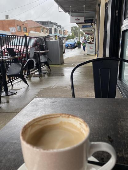 Mug of cappuccino with from already consumed sitting on a dark wooden coffee table. Rainy suburban shopping street in the background. Signs for a barber shop, butcher, physio, solicitor, and a small grocery store. Most of the other cafe tables have been rained on and the chairs tilted to rest on them so the rain drips off.