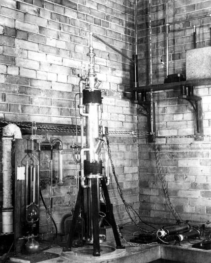 A black and white photograph depicts the first electron microscope in North America, constructed at the University of Toronto during World War II and the Great Depression. The machine features a tall vertical column with multiple glass components, connected to vacuum pumps and wiring systems. It sits on a tripod-like stand against a brick wall. This image provides insight into scientific research during a challenging historical period.