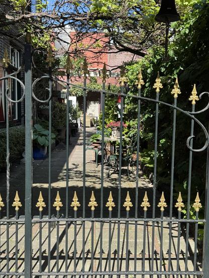 Die Einfahrt zu einem Haus ist durch ein geschlossenes Gartentor mit Schmiede arbeiten und goldenen Pfeilspitzen abgesichert. Auf dem gepflasterten Weg sieht man einen Tischstühle, und weiter hinten Gartenmöbel