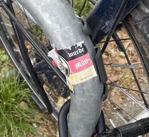 Nazi-Aufkleber auf Fahrradständer. Schwarz-rot-Gold mit Frakturschrift, nicht ganz sichtbar: ein WK2-Soldat mit Stahlhelm