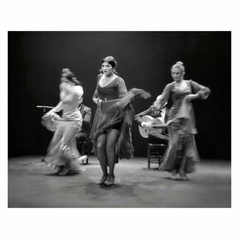 This is a black and white landscape format image of three female flamenco dancers on a theatre stage. The face of the woman in the centre is in focus, and much of the remainder of the image is a whirl of movement. A man seated playing guitar is just visible in the background. The dancers are lit by spotlights and the backdrop is dark, creating a high contrast. Taken with a Sony RX100 in Sevilla.