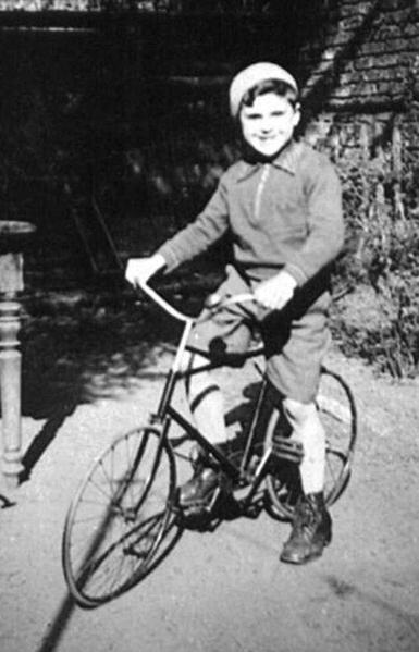A boy photographed outside, in front of a brick building. He is on a bicycle. He is wearing short trousers and a long-sleeve blouse.