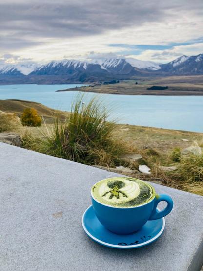 A photo of a matcha latte in a blue mug and saucer on a grey table. The latte art has a silhouette of an alien on it. In the near background are grass and green plants. In the distance is a lake and snowcapped mountains.