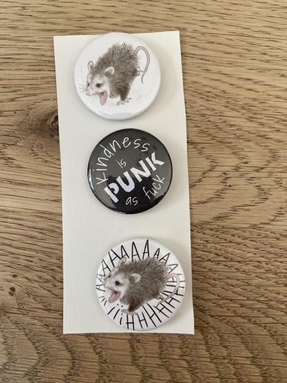 Drie buttons op een papiertje. Eentje van een getekend ratje met zij bekje wijd open. Een zwarte met de tekst “kindness is punk as fuck” en eentje met hetzelfde ratje maar dan met de letters AAAAAAH er omheen.