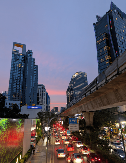 Center of picture shows heavy three lane traffic as cars stuck bumper to bumper. Sun sets in the horizon. View of tall buildings on each side as well as sky train bridge on the right