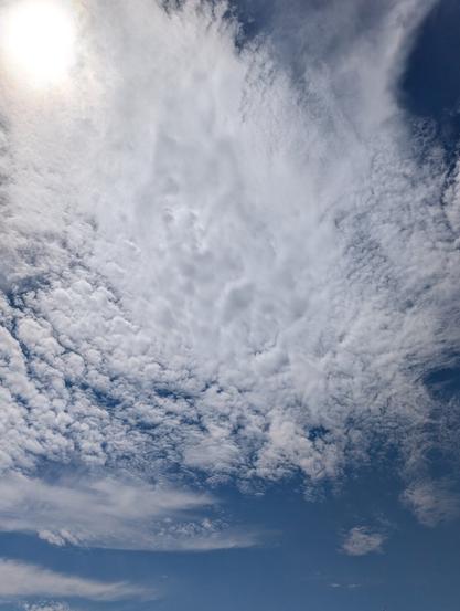 A low-angle, full shot of a blue sky with white, wispy cirrocumulus clouds. The sun shines brightly in the upper-left corner. The clouds cover the majority of the frame, with patches of blue sky peeking through, especially in the lower third of the image.