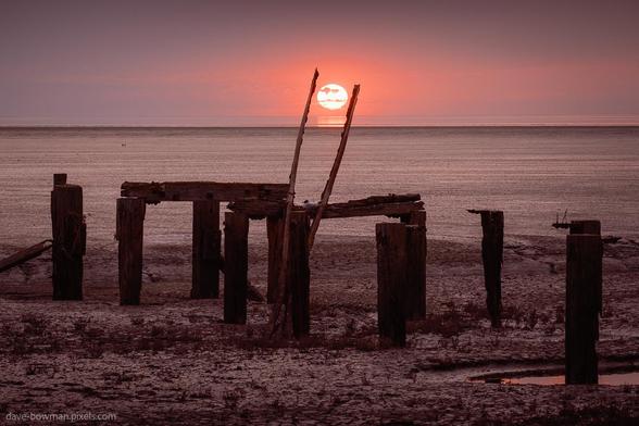 A photograph of an old, decaying wooden jetty at Snettisham in Norfolk, captured as the sun begins to dip toward the horizon behind it. Two upright beams rise from the structure, appearing to reach toward the sun, adding a subtle, playful touch. The weathered timber contrasts with the soft glow of the evening light, creating a calm atmosphere that suggests the quiet passage of time.