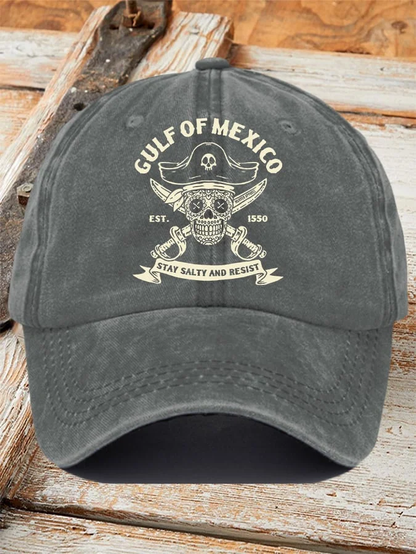 Product photo of a grey baseball cap emblazoned with a pirate style logo featuring a Dia de los Muertos skull, a pirate hat, and crossed swords.  "Gulf of Mexico" is printed across the top, and a banner at the bottom reads "Stay salty and resist."
