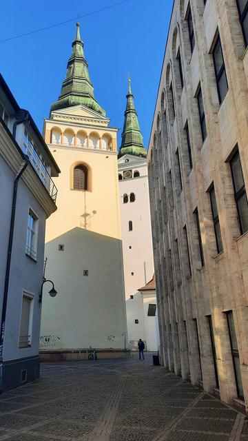 Photo looking down a narrow town pedestrian side-street. There are two tall towers with a square footprint and pointy, top covered by sheet copper oxidized green. On the right side is a four story building with marble facade and many windows. On the opposite side is a lower three story house. The street is paved by cobblestones.