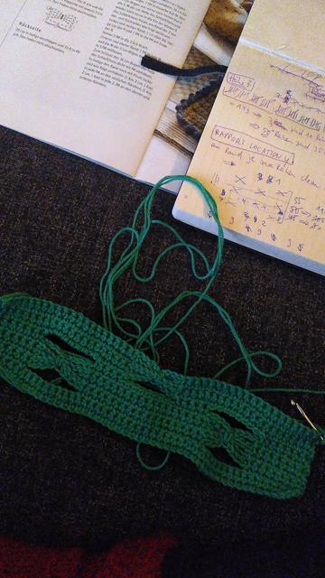 Open books with crochet patterns and my handwritten pattern advancements and calculations, in front of that there's a green crocheted part of a pot holder to be with small "holes" that approximate interwoven crocheted bows