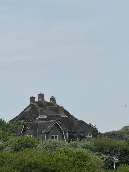 A thatched-roof house is nestled among lush green trees, with multiple chimney stacks visible on its sloped roof, under a cloudy sky.

Ein Reetdach-Haus liegt eingebettet in üppig grüne Bäume, mit mehreren Schornsteinen, die auf seinem schrägen Dach unter einem bewölkten Himmel sichtbar sind.