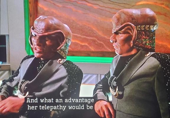 Two Ferengis lean against a bar chatting about Deanna and her mother Lwaxana. The one seen on the left says "And what an advantage her telepathy would be."