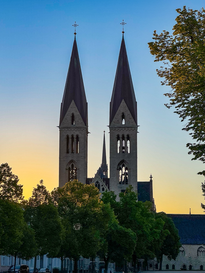 Zwei Kirchtürme mit spitzen Turmhelmen aus Kupfer vor gelborange bis blau eingefärbtem Morgenhimmel, links und rechts stehen die Bäume des Domplatzes