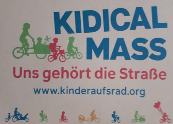 Das Bild zeigt ein Plakat mit dem Titel "KIDICAL MASS" in großen blauen Buchstaben. Darunter steht in roten Buchstaben "Uns gehört die Straße". Die Website "www.kinderaufsrad.org" ist in blauer Schrift angegeben. Das Plakat zeigt Silhouetten von Kindern und Erwachsenen, die auf Fahrrädern fahren. Es gibt verschiedene Farben, darunter Grün, Rot, Blau und Orange, die die Silhouetten darstellen. Die Silhouetten zeigen Familien, die zusammen auf Fahrrädern fahren, einschließlich eines Kindertransporters. Am unteren Rand des Plakats sind weitere Silhouetten von Kindern und Erwachsenen, die auf Fahrrädern fahren, zu sehen. Einige der Silhouetten haben Ballons.