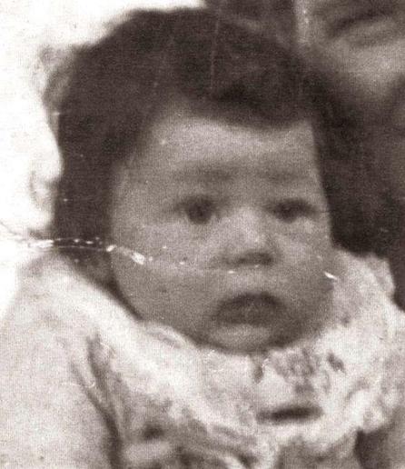 The face of a baby girl with a bib around her neck. She has dark disheveled hair. A fragment of a woman's face can be seen next to her.