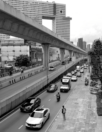 Person standing on the sidewalk waiting for ride to arrive. Photo also shows multi level road, along with sky train and a building that looks like elephant