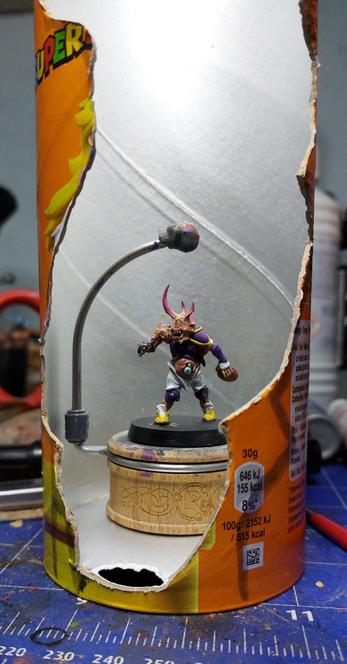 A Pringles can worth a random pattern carved out on one side within it a rathcore miniature holder with a heavily modified blood bowl rotter on an unpainted base. The rotter has elf arms and a plague bearer head and massive eyes growing out of its stomach because this is what you do when you are a rotter.