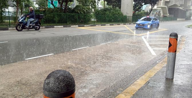 Looking out from a bus bay, onto a Singapore two-lane street during a tropical downpour.  On the left, a motorcyclist braves the weather. On the right, a blue taxi driving in the closest lane, with its parking lights on.  In the foreground, the bus bay, and concrete bollards protecting the pathway beside it.  In the background, the median strip, protected by a green metal fence, filled with tropical plants.  The concrete column holding up the overhead light rail can also be seen behind the fence.