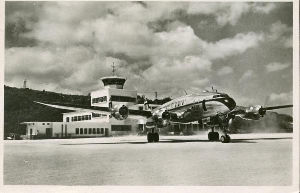 The postcard is black and white. The plane is seen pulling away from the terminal building in the back.