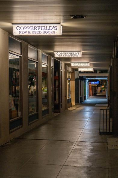 A photograph of a dark night sidewalk in front of a bookstore, primarily illuminated by the small lights for Copperfield's New & Used Books above the walkway.