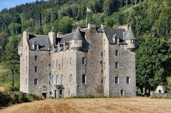 Castle Menzies. The image shows a very large tower house. It is four storeys plus an attic high and has a main block with corner extensions on the far left and near right. At rooftop level is it decorated with small turrets. There is a field in the foreground and a rising slope covered in trees in the background. The scene is in sunshine.