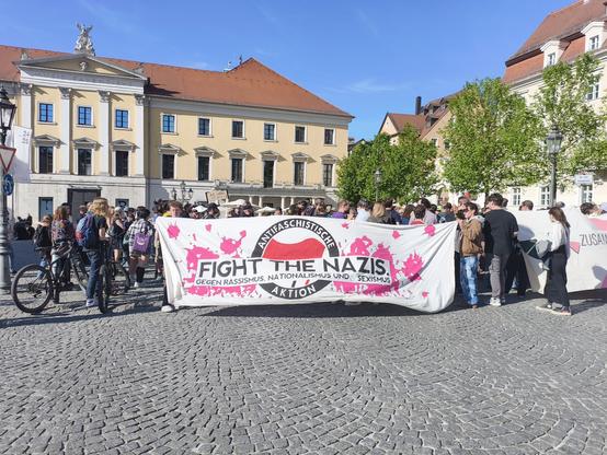 The image depicts a protest in a European city square, with a large crowd gathered. The central focus is a large banner held by the protesters, which reads "FIGHT THE NAZIS" in English, along with the German text "GEGEN RASSISMUS, NATIONALISMUS UND SEXISMUS" (Against racism, nationalism, and sexism). The banner also features a logo with the words "ANTIFASCHISTISCHE AKTION" (Anti-fascist Action) encircling a red circle with a white center, resembling the Nazi symbol, but with a pink splatter. The crowd is diverse, with people of various ages and attire, some holding bicycles. The background shows a classical European building with a yellow facade and red roof, typical of many European cities. The sky is clear and blue, indicating a sunny day. The cobblestone pavement and the architecture suggest a historical setting.

Provided by @altbot, generated privately and locally using Ovis2-8B

🌱 Energy used: 0.254 Wh