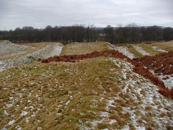 Some of the ramparts of Ardoch Roman Fort. The image shows a view over a rough grassy field with trees in its far side. The ground is indented with a sieres of parallel ditch features running from the bottom of the frame and into it. The pattern of the ditches is made more noticeable by snow that has collected in the hollows. The scene is cloudy.