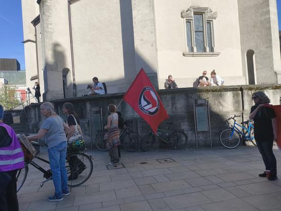 The image depicts a scene in front of a large, light-colored building with a stone facade. A red flag with a circular logo and the text "ANTIFASCISME ACTION" is prominently displayed in the center. Several people are present, some standing and others sitting on a low wall. A person in a purple safety vest is pushing a bicycle, while another individual stands nearby with a bicycle. Bicycles are parked along the wall, and a blue bicycle is visible on the right side. The ground is paved with light-colored tiles. In the background, a person is seated on a step, possibly taking photographs. The sky is clear and blue, indicating a sunny day. The overall atmosphere suggests a gathering or protest.

Provided by @altbot, generated privately and locally using Ovis2-8B

🌱 Energy used: 0.187 Wh