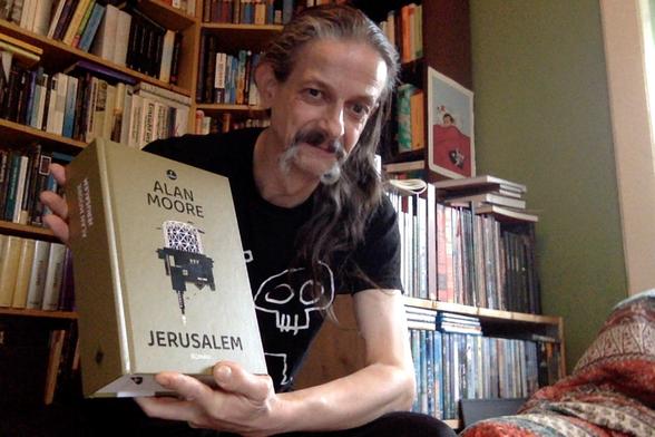 So sieht ein Molo kurz vorm Weinen wegen Eu-Stress aus; mit dickem deutschen »Jerusalem«, frisch aus der Post in den Armen vor meinem Alan-Moore- und Ghibli/Anime-Regal. Im Hintergrund die Sachbuchabteilung meiner Bibliothek.