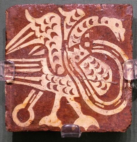 A reddish tile with a cream coloured design of a swan with its wings held up and a crown and chain around its neck.
