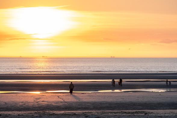 A coastal scene at low tide illuminated by the light of the low-hanging sun, which is visible in the frame. The beach is dotted with puddles that reflect the sunlight, creating a shimmering effect. Silhouettes of several people stand at the shore, gazing out at the sea. On the horizon, the outlines of three ships are visible, their shapes vertically mirrored by the phenomenon of mirage, enhancing the serene beauty of the scene.