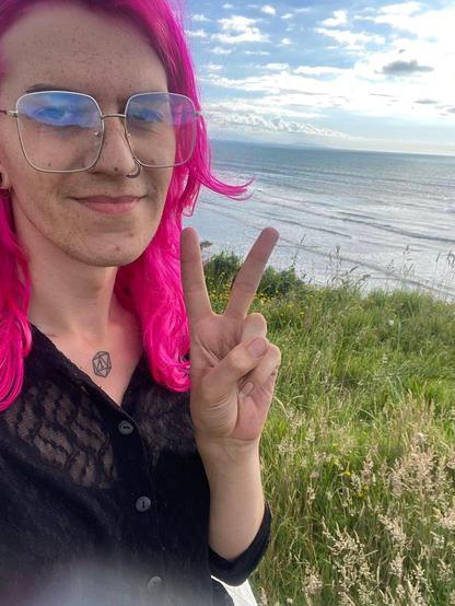 Photo of Lucy, a 30yo light-skinned woman with bright pink hair; she has a tattoo of a 20-sided dice on her clavicle. In reality she has recently died her hair blonde.