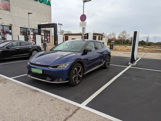 Blue Kia EV6 at a Tesla Supercharger in Tarragona, Spain, with dusty desert-like ground in the background.