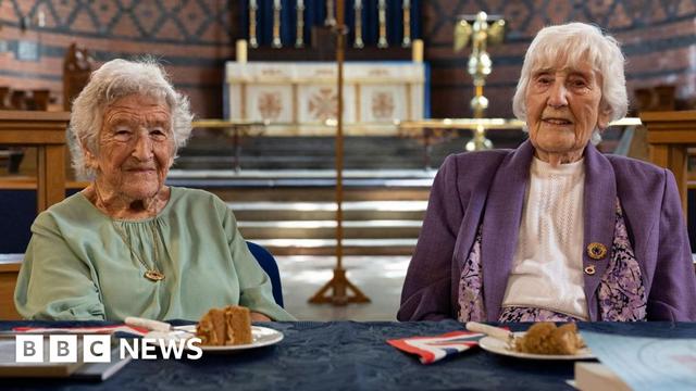 The two codebreaking ladies sitting in the church where they have been presented with an award. Kath is wearing a green top while Gwenfron is wearing a purple jacket, both have short white hair. There is cake laid on the table along with a framed a certificate and a book.