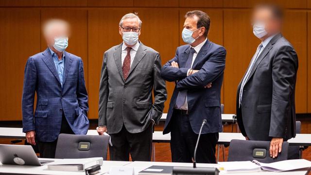 Die vier Angeklagten stehen an Tischen vor einer holzgetäfelten Wand, sie tragen Masken. Zwei Gesichter sind verpixelt.