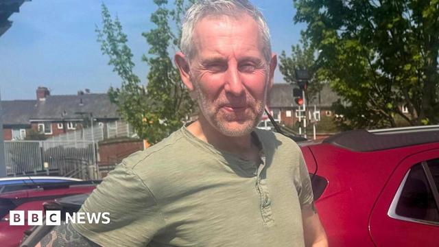 A man with short grey hair and a khaki t-shirt stands in a car park. A man with short grey hair and a khaki t-shirt stands in a car park.