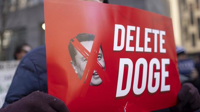 A protestor holds a sign that has a picture of Elon Musk with an X over his face, and text that says "Delete DOGE"