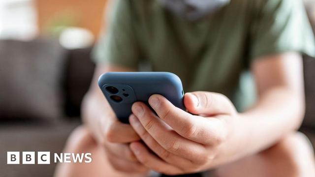 A teenage boy, wearing a dark green t-shirt, is shown blurred, sitting on a sofa. The image is focused on a blue smartphone he is holding in his hands.