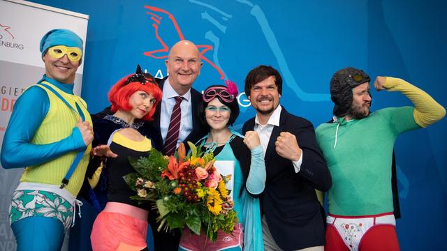 Trashig als Superhelden kostümierte Leute nehmen eine Auszeichnung entgegen und posieren dabei albern.