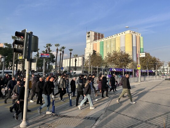approximately 30 people crossing the street, most people are wearing black leather or textile jackets and blue jeans, there’re trees, i.e. palms and presumably a factory in the background 