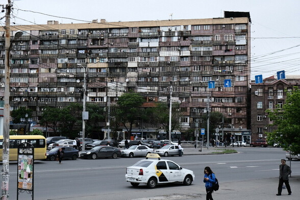 a view on a 11-floor house, which is quite flat and covered with differently decorated balconies. there is also a street in front of the house, where a few cars are parked and one taxi car is driving from the point of view