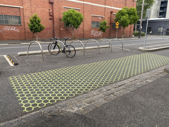 A photo showing the bike parking bays that are spaced out all along Wellington Street in Melbourne. Just a cool idea to take a few car parking bays all along the street and fit them with bike parking U-rails. Then even cooler: they have painted the bike lane with highly contrasting paint in a sort of “net” pattern, which warms bike riders to slow down as well as warning any cars on side streets that they are about to cross a bike lane to merge onto Wellington Street car lanes. 