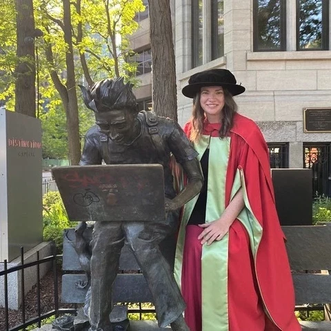 Me sitting in PhD academic regalia next to a statue of a student with disheveled hair hunched over a computer located on the road in front of McGill campus.