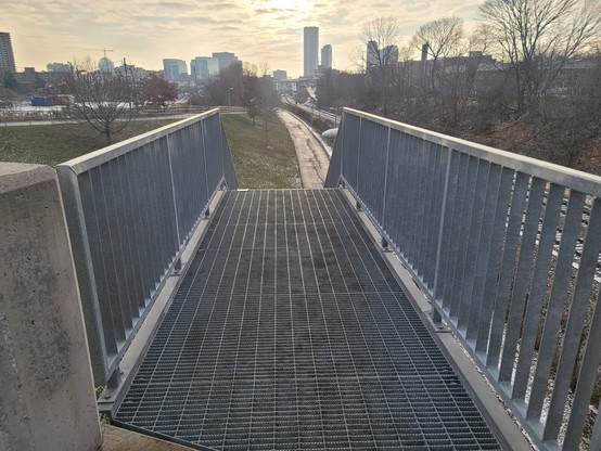 Metal grid platform woth metal railings connecting the top of a set of metal stairs to the sidewalk at the top of a bridge.