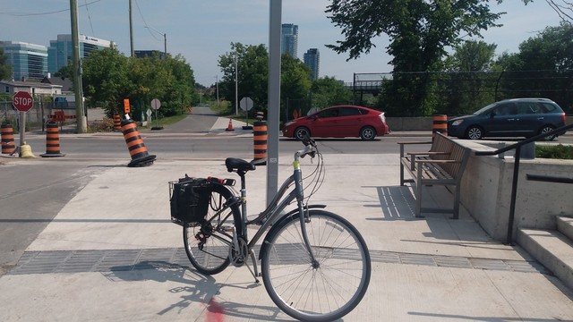 Silver step through bike standing on fresh sidewalk paving in front of Gladstone. Trillium pathway continues across the road