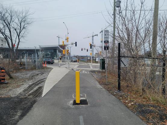 Multi use pathway apparoaching a signalized intersection with a yellow bollard in the middle and a small section of sidewalk on the left. On the right, a signpost with "bikes yield to pedestrians" sign and "peds keep left, bikes keep right" sign.