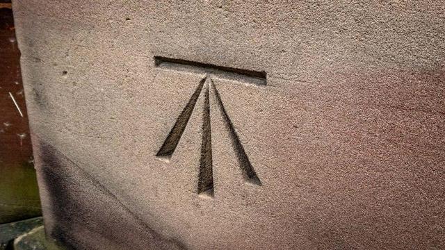 A non-print example.

The most common form of the symbol, although other variants exist. It's carved into a smooth block of stone on the side of a building or monument plinth, and it looks like a capital T with an upside down V overlaid onto it so that the two angled lines from the V come together with the T's vertical stroke to meat its horizontal stroke at a single point. All the bottom lines taper toward that point.  Looking at it another way, it's a horizontal line with an arrow pointing at it, saying "here, this level!"

They were used for marking height reference points during various surveys of the British isles.

Photo by Mike Taylor on geograph.org.uk, CC:by
