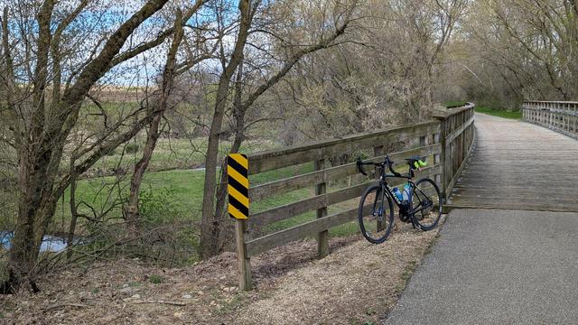 Blue and black Roubaix bicycle leaning on the wooden railing of a wooden decked bridge over the North Fork of the Whitewater River on the Great River Ridge State Trail looking North. The river can be seen to the left weaving through pasture with black cows grazing.