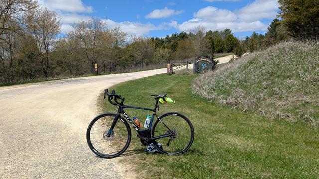 Blue and black Roubaix bicycle parked off the gravel entrance road to Carley State Park with brown gates with signs and the wooden decorated "Carley State Park" sign with an image of Virginia Bluebells in bloom. The road curves to the right and there are trees just leafing out on either side.